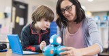 A female teacher sits next to a first grade boy and helps show him how to program a robot using a digital tablet.